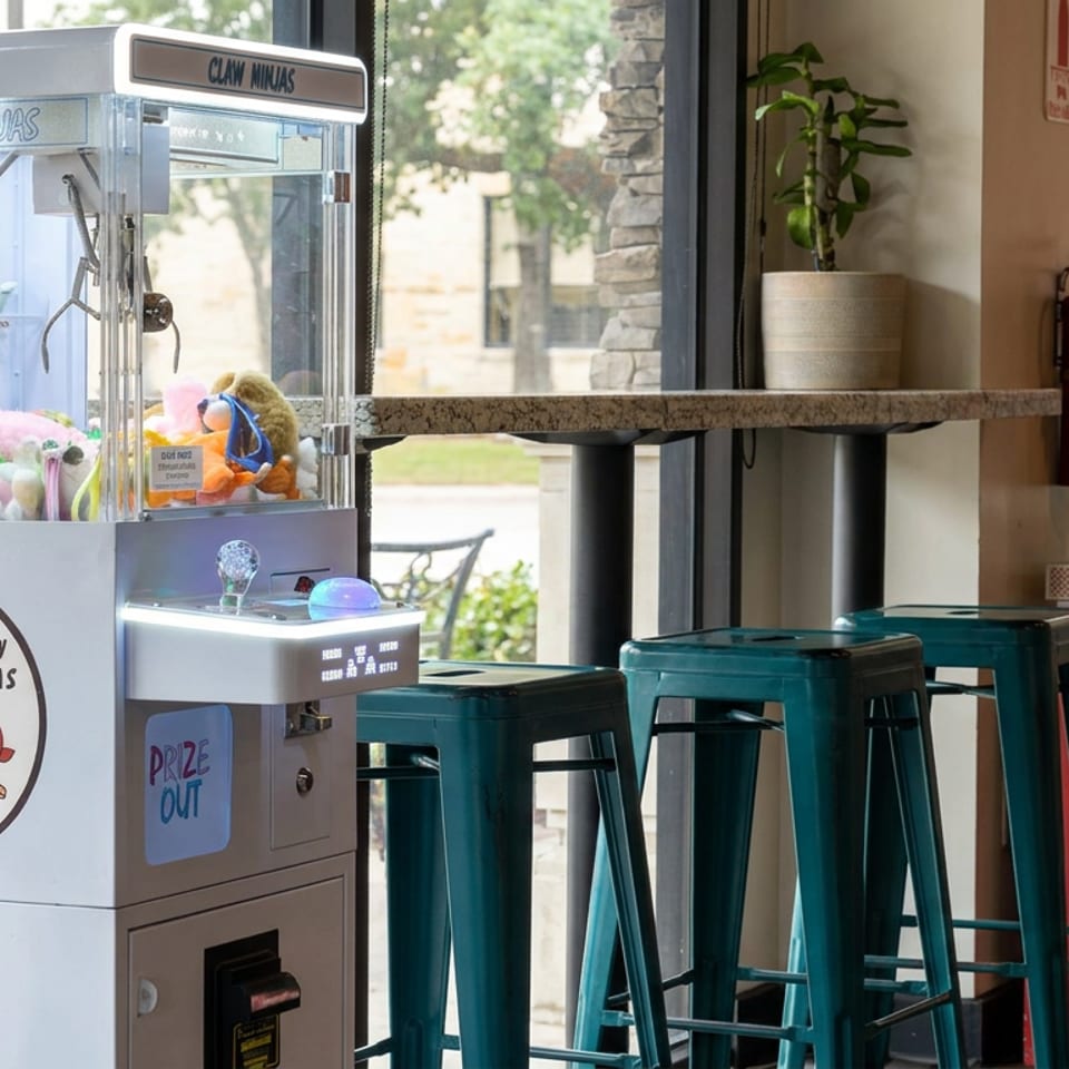 Claw Ninjas claw machine filled with plush toys beside teal bar stools, a counter, potted plants, and board games.