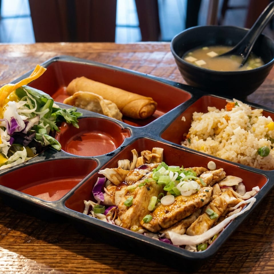 Bento box with grilled chicken, fried spring rolls, fried rice, salad, and miso soup on a wooden table.