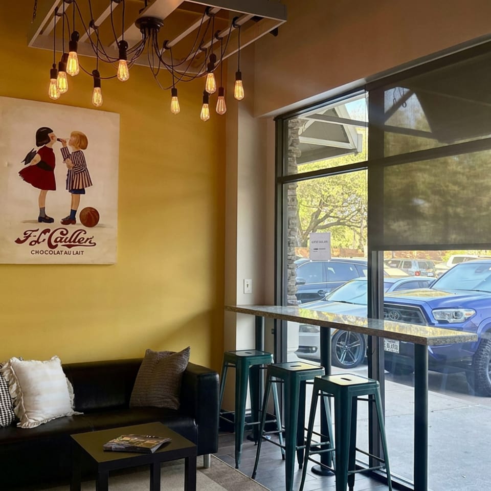 Cozy café seating area with black sofa, pillows, high tables, pendant lights, and vintage chocolate advertisement beside large windows.