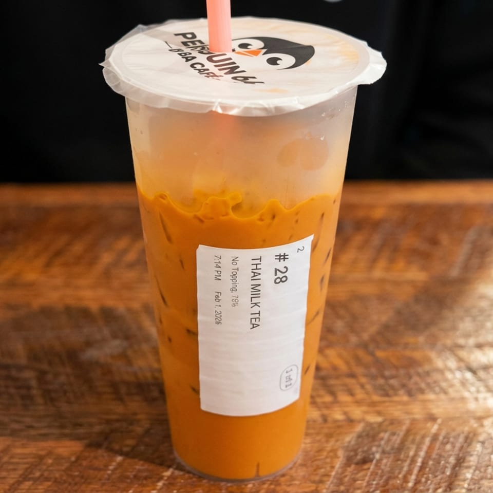 Iced Thai milk tea in a branded plastic cup with straw, sitting on a warmly lit wooden table.